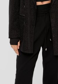 Zwarte pailletten blazer met een ontspannen pasvorm en twee voorzakken. Vergezeld van zwarte elastische taillebroek. Gladde textuur, glanzende accenten.