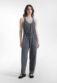 Mono de mezclilla gris con tirantes ajustables, cintura elástica, cordón y dos bolsillos delanteros. Textura suave y ajuste relajado.