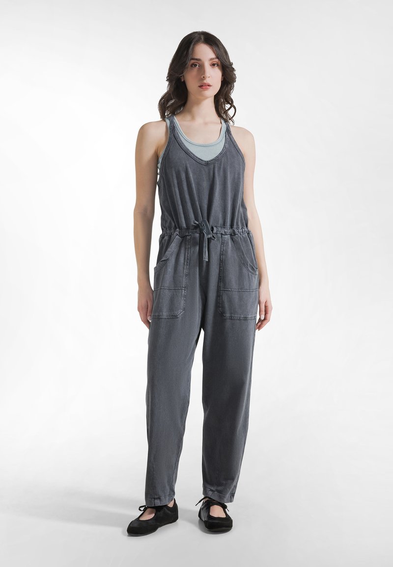 Mono de mezclilla gris con tirantes ajustables, cintura elástica, cordón y dos bolsillos delanteros. Textura suave y ajuste relajado.