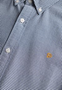 Camisa de cuadros azul y blanca, hecha de tela, con un cuello abotonado, botones blancos y un pequeño logo amarillo.