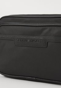 Schwarze Nylon-Tommy-Hilfiger-Tasche mit strukturierten Oberflächen, die mit einer auffälligen Logo-Platte und kontrastierenden Nähten entlang des Reißverschlusses ausgestattet ist.