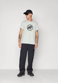 Lichtgrijs T-shirt met een zwarte mammoetafbeelding in het midden, gecombineerd met donkere cargobroeken en zwarte schoenen, gedragen door een persoon.