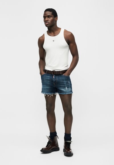 Νέος άντρας που φοράει άσπρο αμάνικο μπλουζάκι, ξεβαμμένα τζιν σορτς με ζώνη, σκούρες κάλτσες και καφέ μποτάκια με κορδόνια, στέκεται με τα χέρια στις τσέπες.
