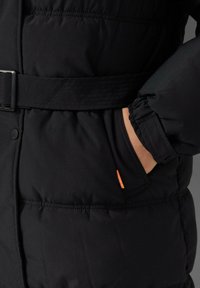 Crna puffer jakna s pojasom u struku, elastičnim rukavima i bočnom džepom s malom narančastom etiketom. Glatka tekstura, kviltani dizajn.