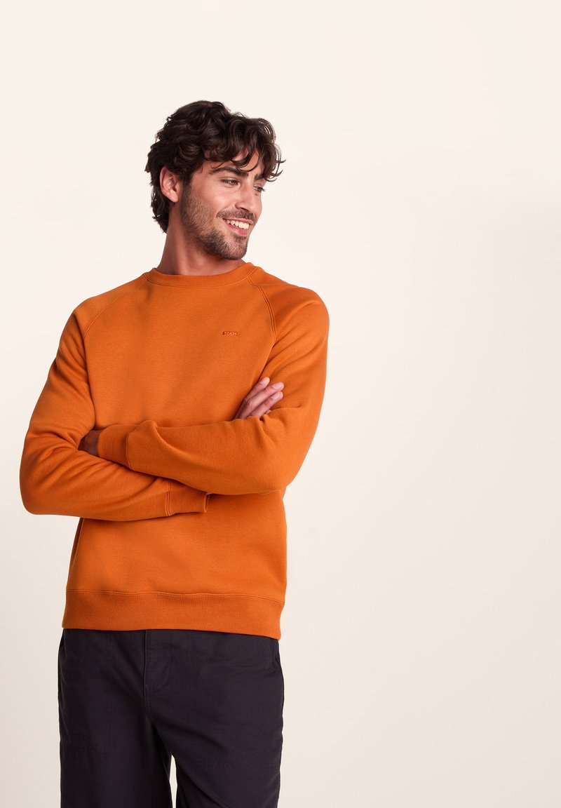 Man with curly hair wearing an orange long-sleeve sweatshirt and black pants, arms crossed, smiling, looking to the side against beige background.