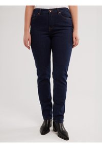 Persona, kas valkā tumši zilas pieguļošas džinsa bikses un melnas potīšu zābakus, stāvot pret vienkrāsainu gaišu fonu.