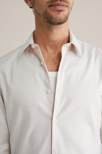 Hvid skjorte med knapper lavet af glat stof, med en klassisk krave og diskret tekstur, delvist knappet op over en almindelig hvid undertrøje.