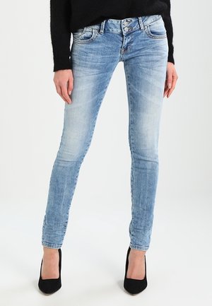 Frau trägt hellblaue Skinny-Jeans mit Knopfleiste, schwarzen Pullover und schwarze High Heels, steht mit entspannten Händen seitlich.