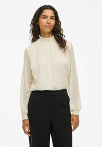 Cream long-sleeve blouse with chevron pattern, high ruffled collar, and elastic cuffs, paired with black tailored trousers.