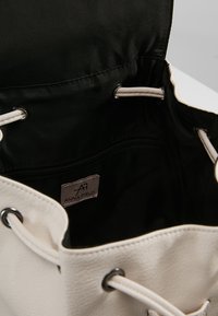 Bolso tipo bucket con un exterior de textura crema, forro negro, herrajes de acero inoxidable, cierre ajustable con cordón y un bolsillo interno con cremallera.