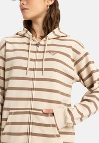Sudadera con cremallera a rayas en color beige con bandas marrones. Presenta una capucha con cordón, bolsillos frontales y un pequeño logotipo en el pecho. Textura suave.