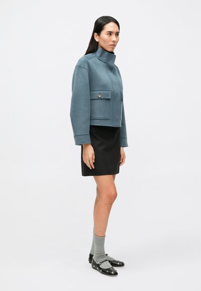 Przycięta niebieska wełniana kurtka z wysokim kołnierzem, dwoma przednimi kieszeniami i detalami w postaci guzików, zestawiona z czarną spódnicą i szarymi skarpetkami.