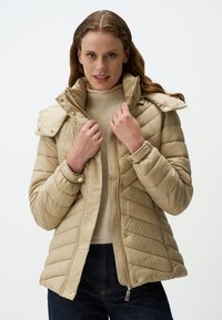 Beige gewatteerde jas met een hoge kraag, drukknoopjes en diagonale stikken. Draag het over een crèmekleurige geribde trui en donkere denim.