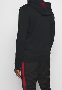 Hoodie noir en tissu doux, doté d'une capuche et d'un accent rouge au dos. Porté avec un pantalon noir présentant une bande latérale rouge.