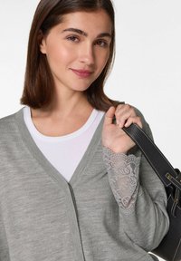 Hellgrauer Cardigan mit V-Ausschnitt und Spitzenakzenten an den Ärmeln, über einem weißen Hemd. Schwarze Handtasche, die von der Person gehalten wird.