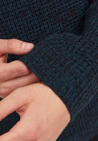 Stickad tröja i mörkblått med strukturerat mönster. Närbild av ärmslutet, som visar en ribbad design och kontrasterande ljusblå accenter.