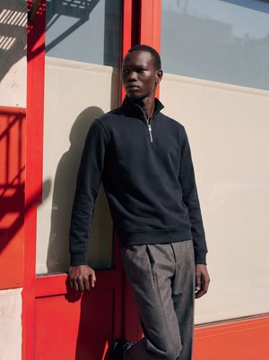 Homme portant un pull zippé noir et un pantalon gris s'appuie contre une fenêtre à cadre rouge lors d'une journée ensoleillée.