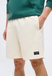 Άνδρας που φοράει σορτς εκρού με πλευρικές τσέπες και μαύρη ετικέτα Björn Borg, συνδυασμένα με πράσινο φούτερ.