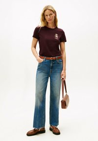 Bordauxfarbendes Baumwoll-T-Shirt mit einem kleinen Grafik, hochgeschnittene, ausgewaschene blaue Jeans mit fransigem Saum, braune Lederloafers und eine braune Handtasche.