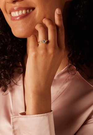Bague en or ornée d'une pierre précieuse transparente ronde, avec un anneau lisse et des accents texturés, portée sur une main contre un chemisier rose pâle.