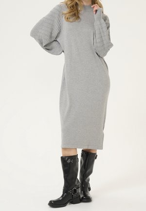 Kobieta w szarej sukience midi z dużymi prążkowanymi rękawami i czarnych skórzanych kozaczkach do połowy łydki na białym tle.