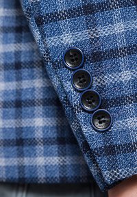 Niebieska marynarka w kratę z teksturowanej tkaniny, z czterema czarnymi guzikami na mankietach. Wzór obejmuje różne odcienie niebieskiego i białego.