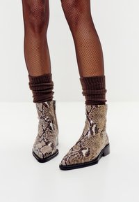Botas de tobillo de piel de serpiente con punteras afiladas, patrones brillantes en marrón y beige, y tacones bajos en bloque, combinadas con calcetines marrones con relieve.