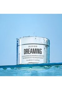 Color Wow Dreaming-behandling i en klar, rund burk med silverdetaljer. Svarta textetiketter visar ingredienser och volym, omgiven av vattendroppar.