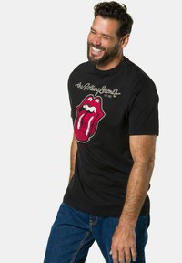 Schwarzes Baumwoll-T-Shirt mit einem abgenutzten roten Lippenlogo, darüber in Grau der Text "the Rolling Stones", Rundhalsausschnitt, kurze Ärmel.