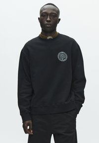 Sweatshirt noir avec un emblème graphique circulaire sur la poitrine, fabriqué en tissu doux avec une coupe décontractée et des poignets et un ourlet côtelés.