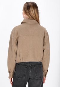 Camicia beige tagliata, con colletto e maniche lunghe, caratterizzata da un tessuto a maglia con una leggera trama. La vista posteriore mostra le cuciture delle spalle e una leggera arricciatura.