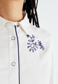 Hvit skjorte med knapper som har blå blomsterbroderi på brystet, kontrasterende blå kant langs knappestolpen, og klassisk krage.