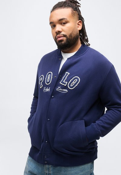 Άνδρας με γένια και κοτσίδες ντυμένος με μπλε σκούρο μπουφάν με κουμπιά και λογότυπο "POLO Ralph Lauren", με τα χέρια στις τσέπες και ανοιχτόχρωμο τζιν.