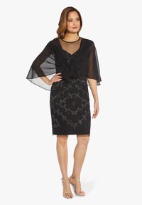 Donna con un vestito nero alla lunghezza del ginocchio, decorato con perline argento e una mantella nera trasparente annodata davanti, posa con una mano sulla vita.