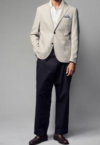Lys beige linned blazer med to frontlommer, parret med mørkeblå bukser og brune loafers med guld accenter.