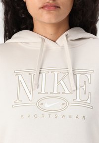 Nike kapota palaidinė kreminės spalvos su dideliu smėlio spalvos logotipu. Su raišteliais ir minkšta audinio tekstūra. Skirta sportinei aprangai.