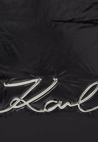 Černý materiál s prošívaným designem a lesklým povrchem, na kterém se nachází vyvýšené metalické stříbrné logo "Karl". Texturovaný povrch s prošívanými detaily.