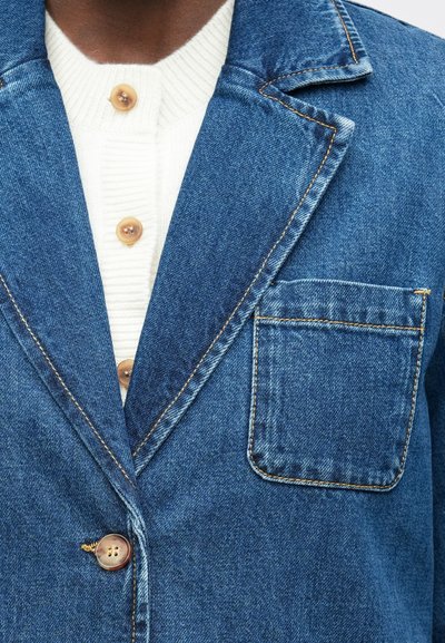 Denim bliuzonas su siuvinėta kišenė, dėvimas ant baltos raštuotos megztinės su šviesiai rudos spalvos mygtukais, matomas viršutinėje krūtinės ir peties zonoje.