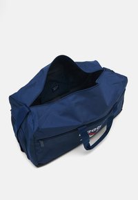 Saco de viagem azul-marinho feito de tecido durável, com fecho de correr, duas alças e um logótipo visível na lateral. O interior é forrado.