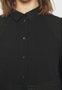 Camicia nera con bottoni, caratterizzata da un tessuto testurizzato, colletto classico e sei bottoni neri sulla parte anteriore. Materiale leggero e semi-trasparente.