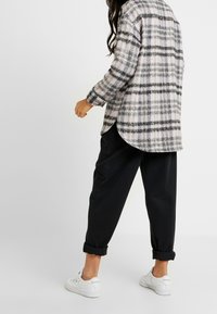 Plaid flannel shirt in beige, black, and gray tones, featuring long sleeves, a curved hem, and soft texture; paired with black trousers and white sneakers.