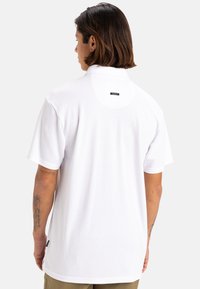 Weißes Polo-Shirt aus leicht strukturiertem Stoff, mit kurzen Ärmeln und einem Etikett auf der Rückseite. Gerader Saum mit Seitenlitzungen für bessere Bewegungsfreiheit.