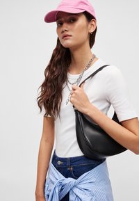 Μαύρη δερμάτινη τσάντα ώμου με καμπυλωτό σχήμα, φορεμένη πάνω από ένα λευκό μπλουζάκι, συνδυασμένη με ένα μπλε ριγέ πουκάμισο δεμένο στη μέση.