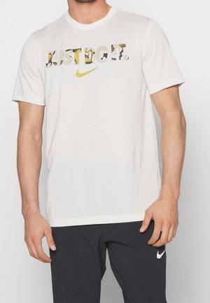 Mężczyzna ubrany w białą koszulkę Nike z złotym napisem "JUST DO IT." oraz czarne spodnie Nike z białym logo na lewym udzie.