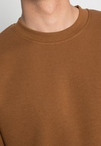 Felpa in cotone marrone con scollo a girocollo, caratterizzata da una consistenza uniforme e cuciture visibili lungo il collo e l'orlo.