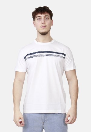 Giovane uomo di fronte che indossa una maglietta bianca a maniche corte con un motivo a pennellate orizzontali scure sul petto e jeans azzurri.