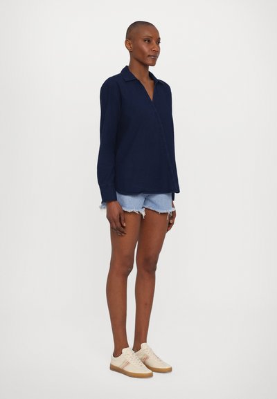 Mörkblå långärmad skjorta med krage, ihopparad med ljusblå denimshorts och krämfärgade sneakers med färgade ränder på sidan.