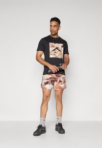 Schwarzes T-Shirt mit kurzen Ärmeln und grafischem Druck, kombiniert mit mehrfarbigen Shorts mit Muster. Das Modell hält eine schwarze Wasserflasche, trägt graue Socken und schwarze Schuhe.