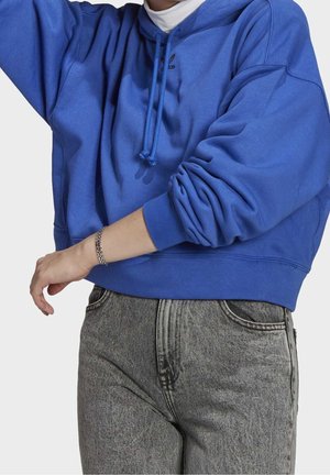 Osoba nosząca niebieską bluzę z kapturem Adidas z podwiniętymi rękawami, srebrną bransoletkę, szare jeansy z wysokim stanem i białą wewnętrzną koszulkę.