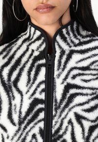 Crna i bijela jakna od flisa s uzorkom zebrastih pruga, visokim ovratnikom i zatvaranjem na zip; ima teksturiranu tkaninu s mekanim osjećajem.